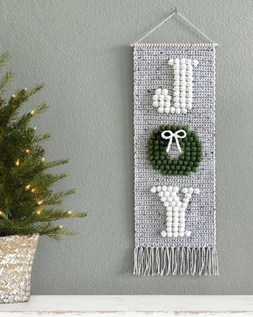 grey crochet wallhanging with white lettering that says "joy", with the letter "o" being a green bobble stitch wreath