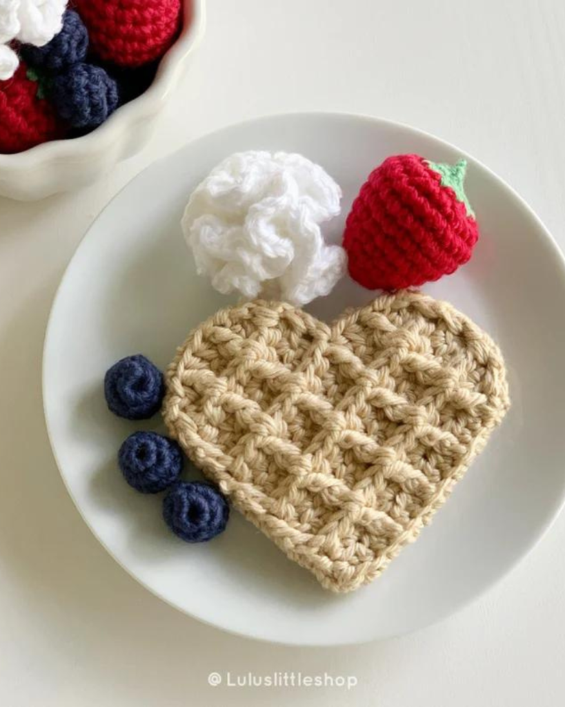 crochet heart-shaped waffle with amigurumi strawberry, blueberries, and whipped cream on white plate