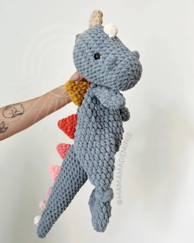hand holding crochet snuggler dino with rainbow spikes