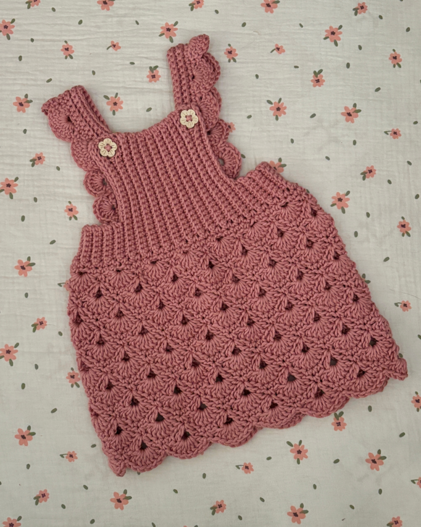 Pink shell stitch crochet baby overall dress with button straps and textured ribbed bib for newborns and infants.
