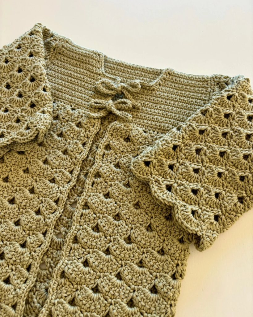 Crochet baby cardigan with delicate shell stitches and two decorative bows on the front.